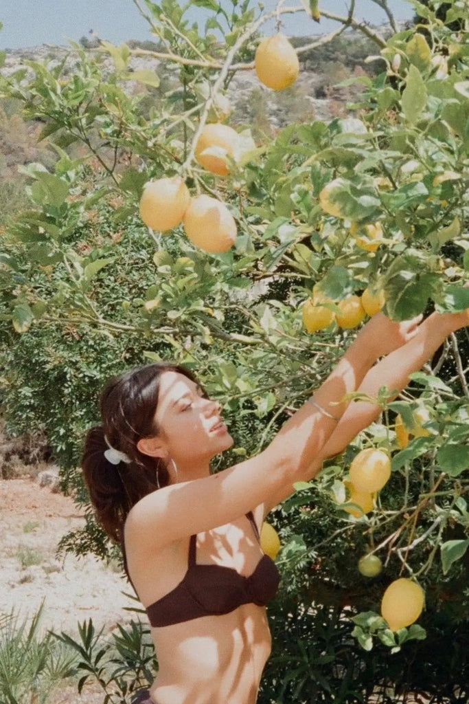 Woman in a bikini standing next to a lemon tree with lemons hanging from it.